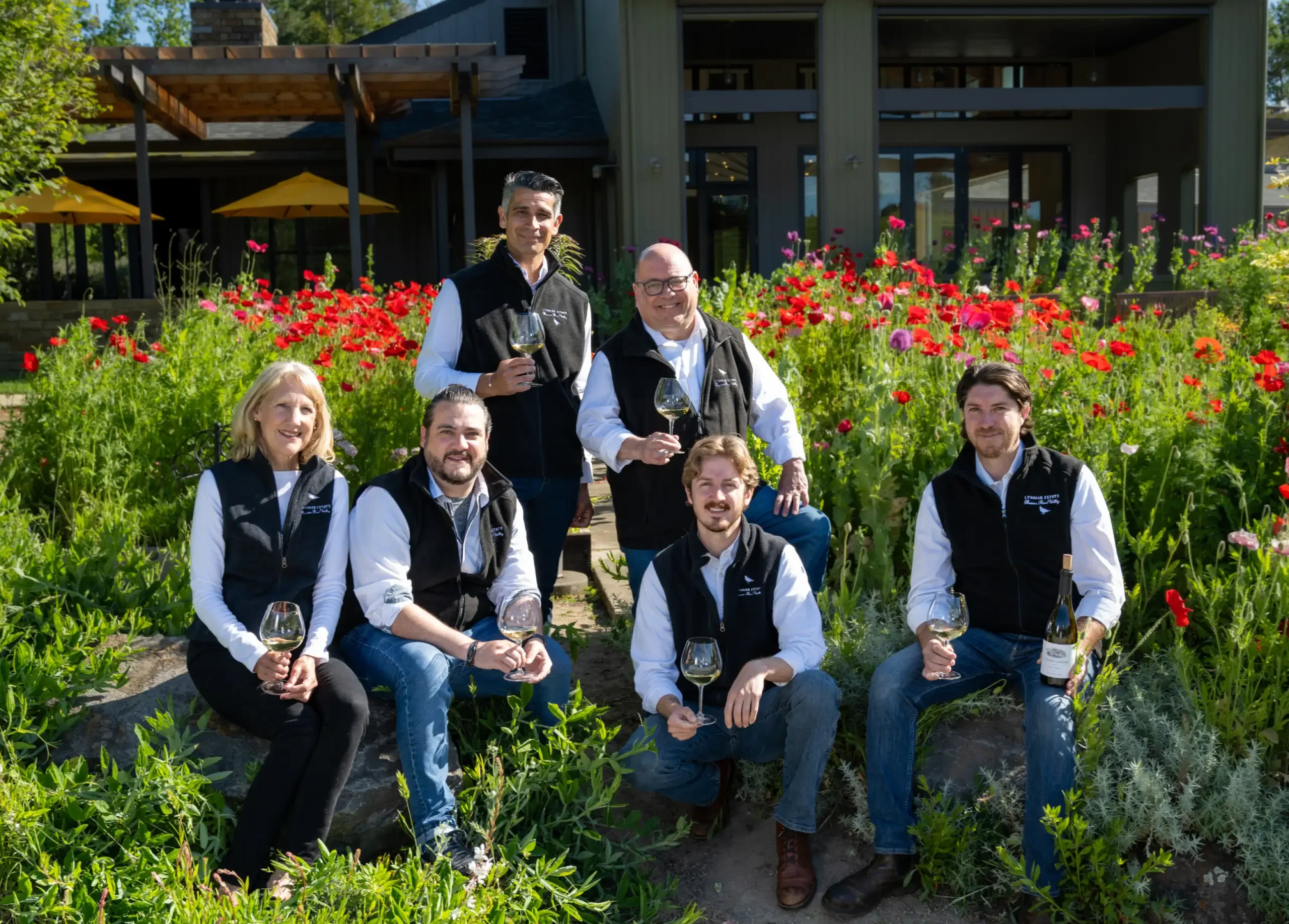 Six adults wearing matching vests pose outdoors in front of a building and a garden with red flowers, each holding a glass of white wine, enjoying a Sonoma wine tasting.