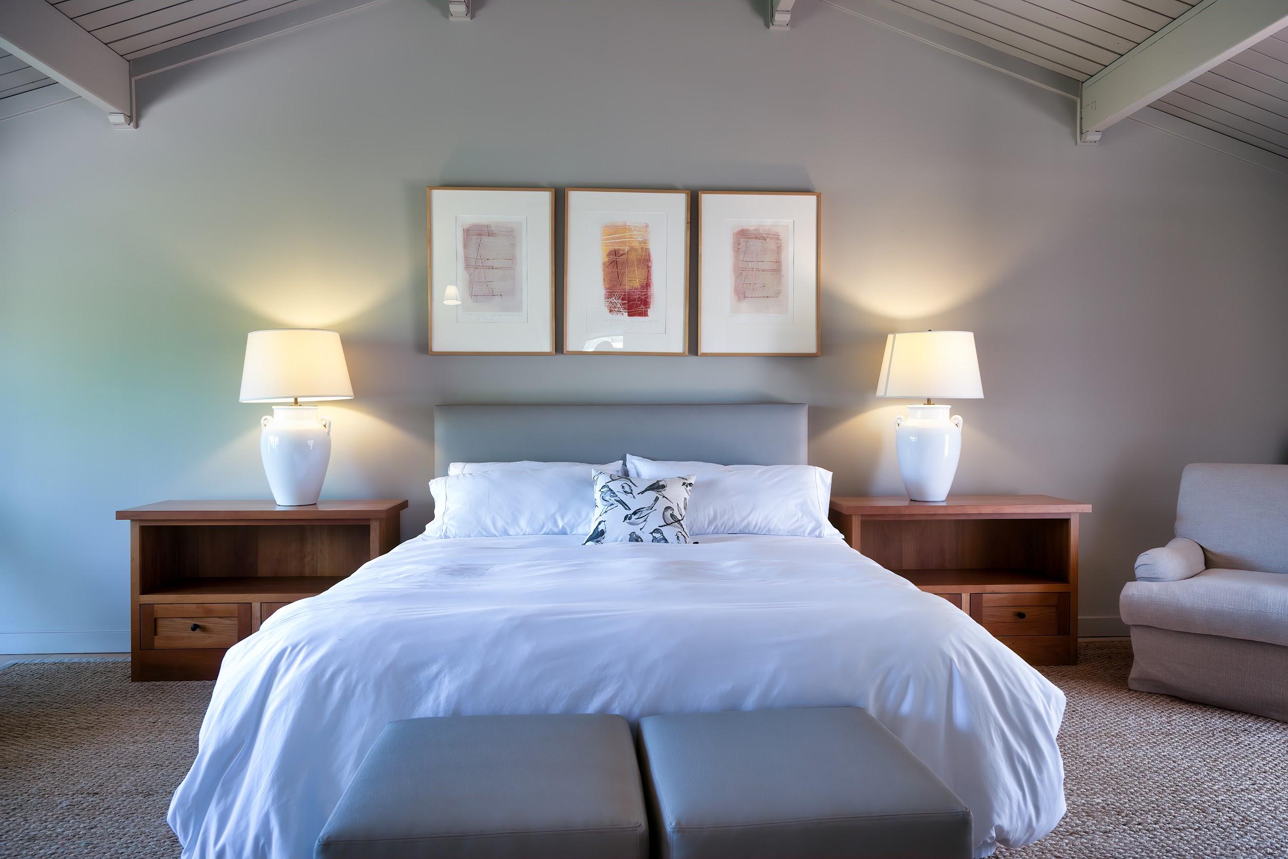 A neatly made bed with white linens is centered between two wooden nightstands, each with a white lamp. Three abstract artworks hang above the bed on a light gray wall—an inviting retreat after an afternoon of wine tasting or savoring Pinot Noir.