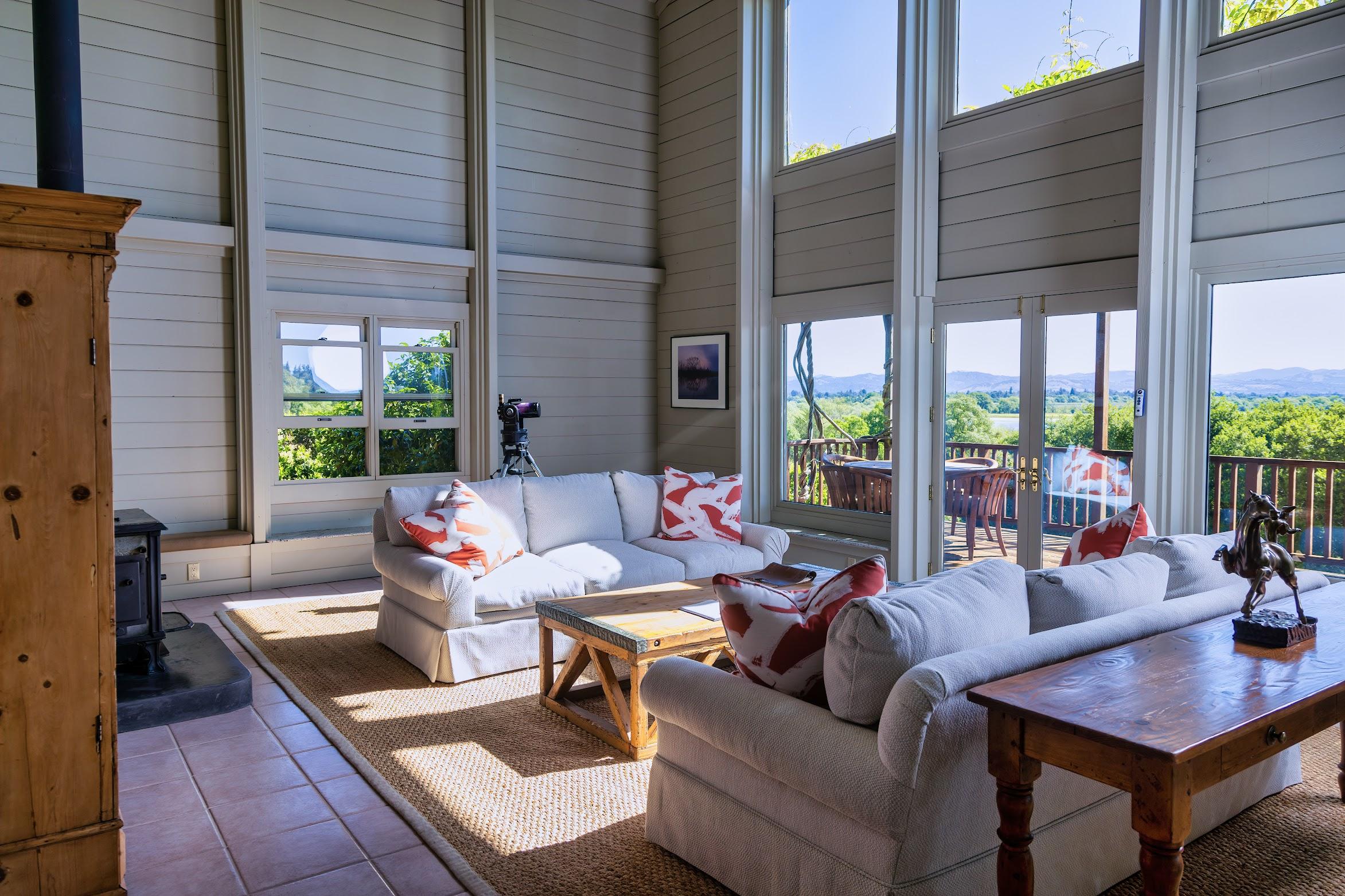 Bright living room with large windows, two white sofas with red-patterned pillows, a wooden coffee table, and a view of a deck and Sonoma landscape outside—perfect for relaxing with a glass of Chardonnay.