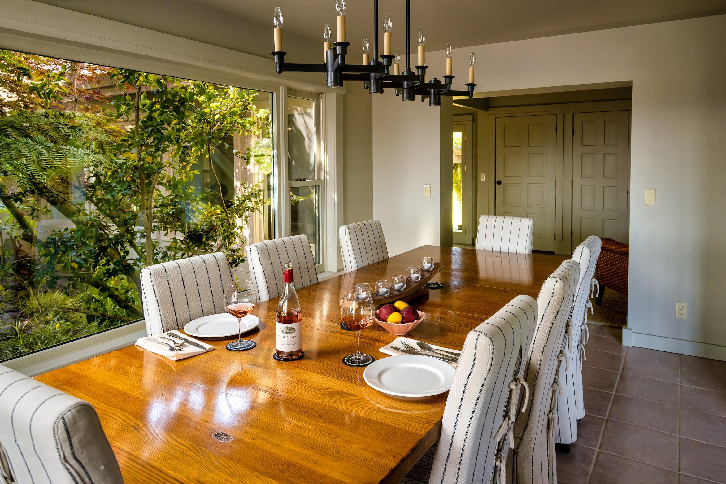 A wooden dining table set for three with wine, plates, and napkins evokes a Sonoma wine tasting experience, surrounded by upholstered chairs in a room with large windows and a modern chandelier.