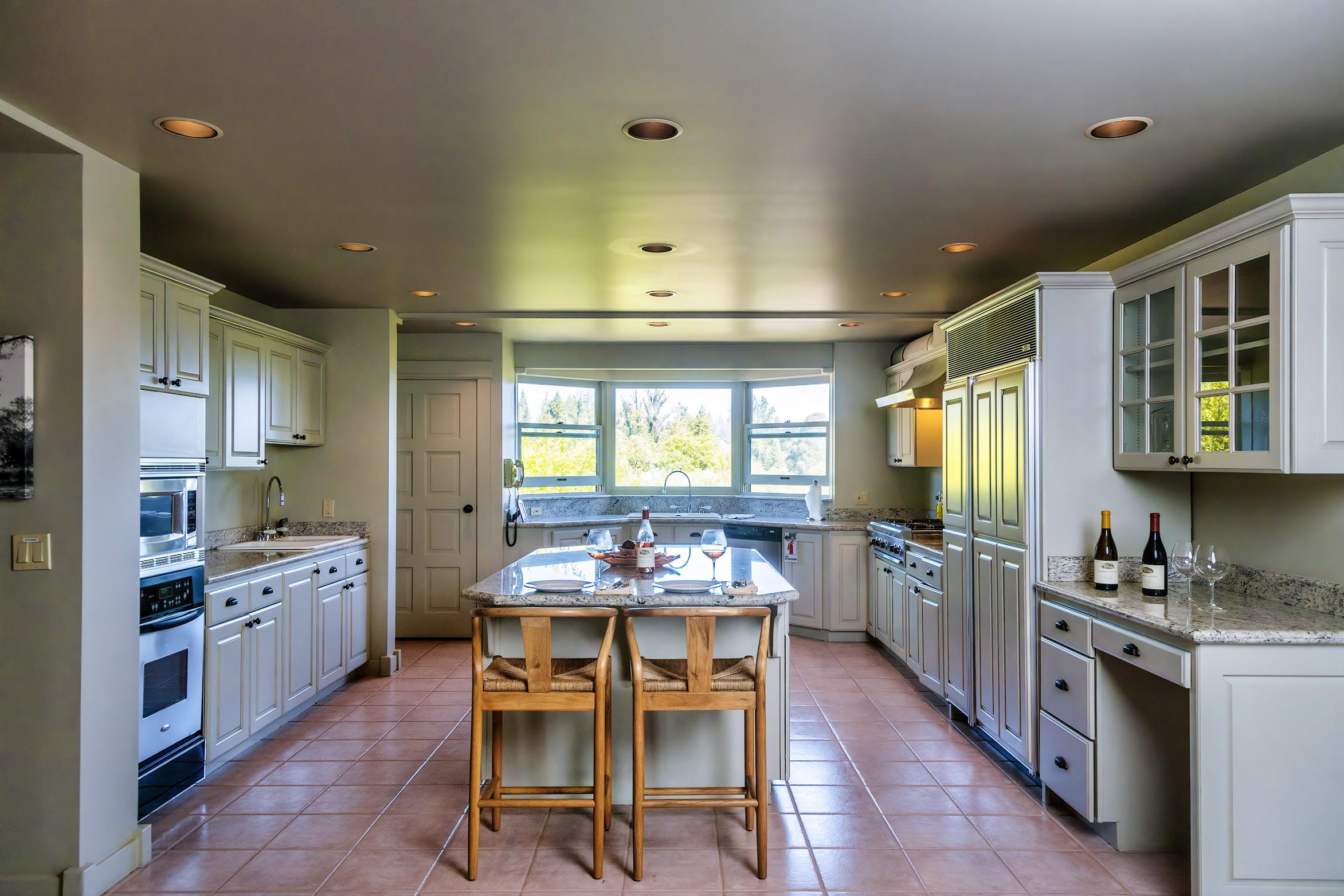 Spacious kitchen with white cabinets, granite countertops, a central island with two chairs—perfect for lunch—with built-in appliances and large windows overlooking the lush greenery of Sonoma’s Russian River.