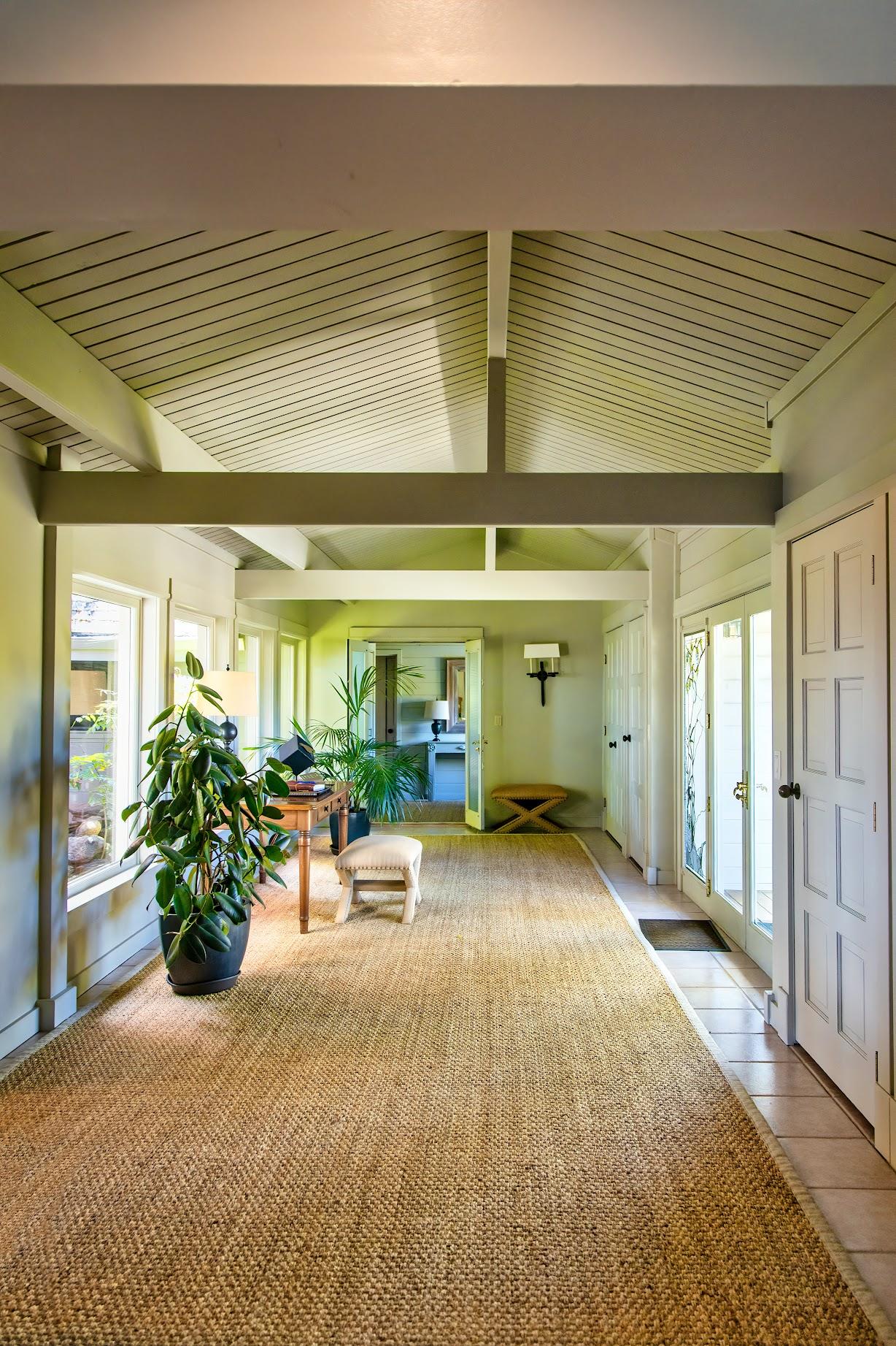 A hallway with high, slanted ceilings, large windows, a woven rug, potted plants, and a bench, leading to an open doorway—perfect for enjoying lunch and a glass of Russian River Chardonnay in the nearby room.
