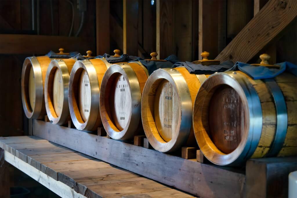 A row of wooden barrels with metal bands sits on a wooden shelf inside a rustic, dimly lit room, aging Sonoma Pinot Noir to perfection.