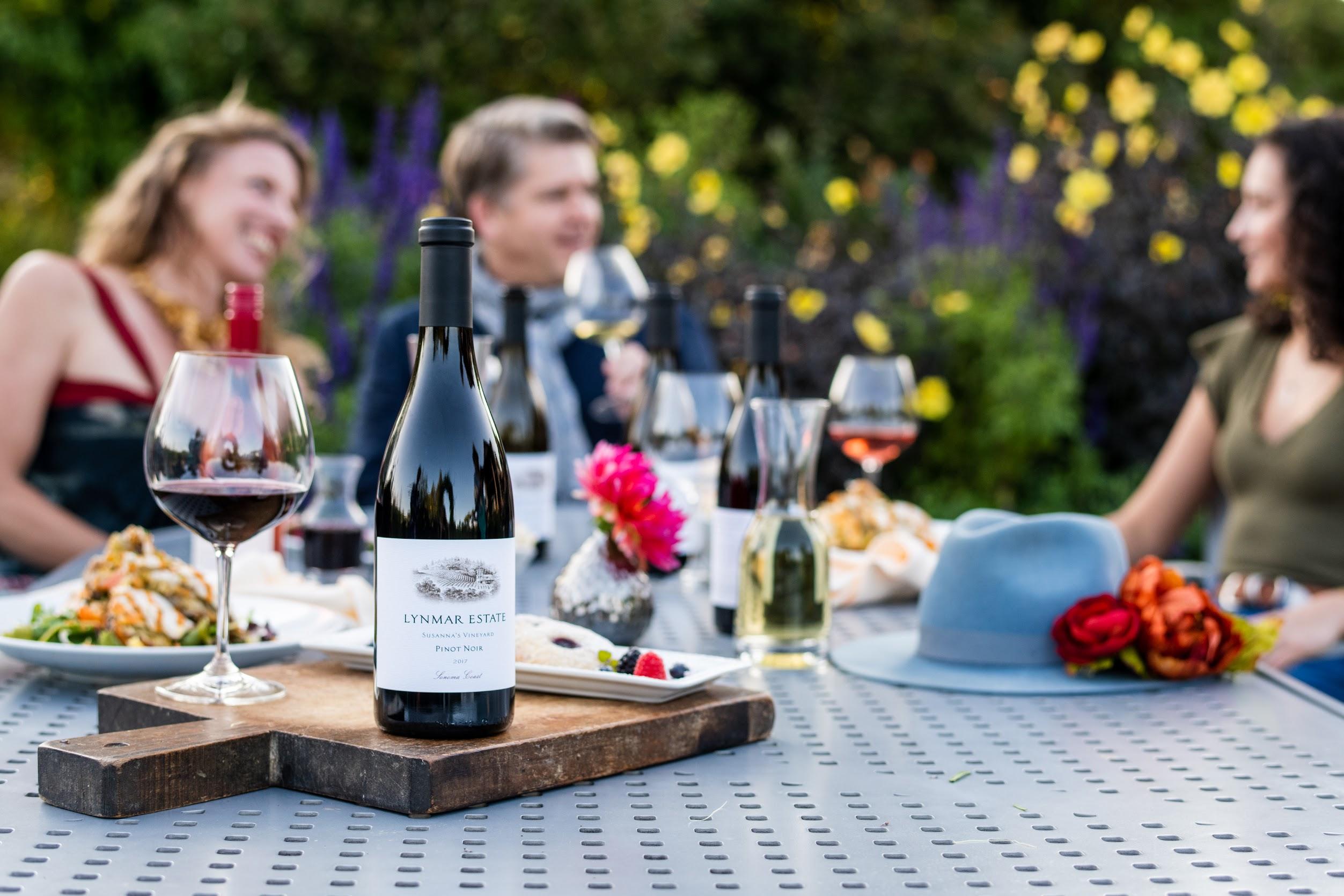 A bottle of Lynmar Estate Pinot Noir and a filled wine glass sit on a table with lunch, food, and flowers, while four people converse in the background during an outdoor wine tasting.