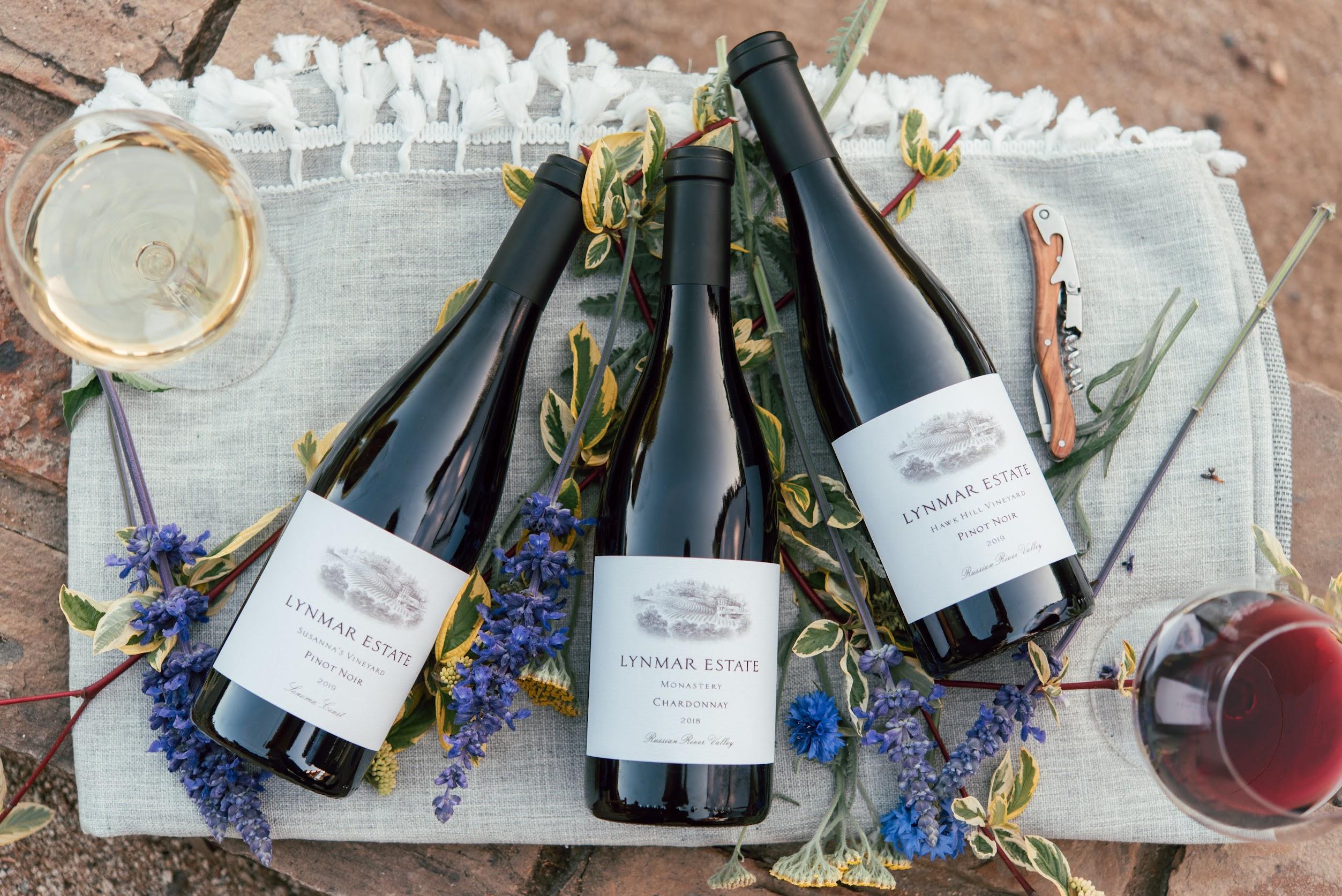 Three Lynmar Estate wine bottles lie on a cloth with flowers, inviting a Sonoma wine tasting experience beside a corkscrew and two glasses—one white, one red—set on a rustic stone surface, perfect for an elegant lunch.