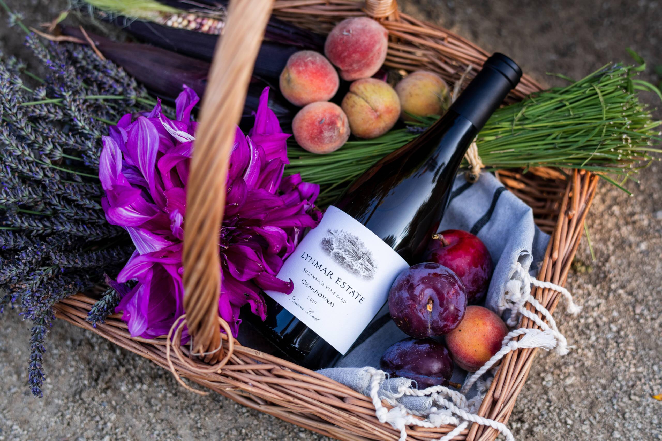 A wicker basket contains a bottle of wine, purple flowers, peaches, plums, purple beans, and sprigs of lavender and green herbs, resting on a light cloth.