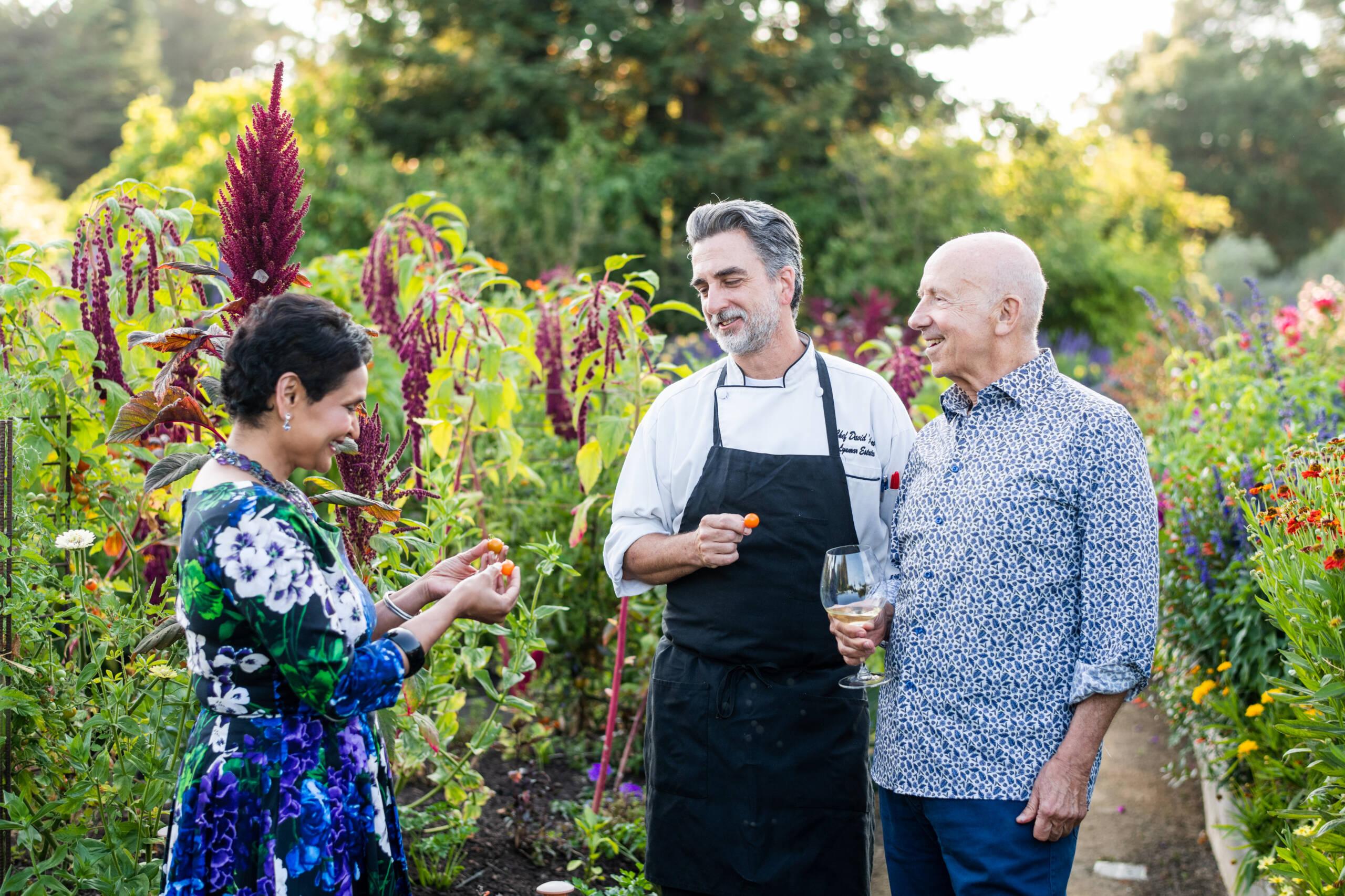 Three adults stand and talk in a lush garden; one wears a chef's uniform, another holds a wine glass, and the third examines a small fruit or vegetable.