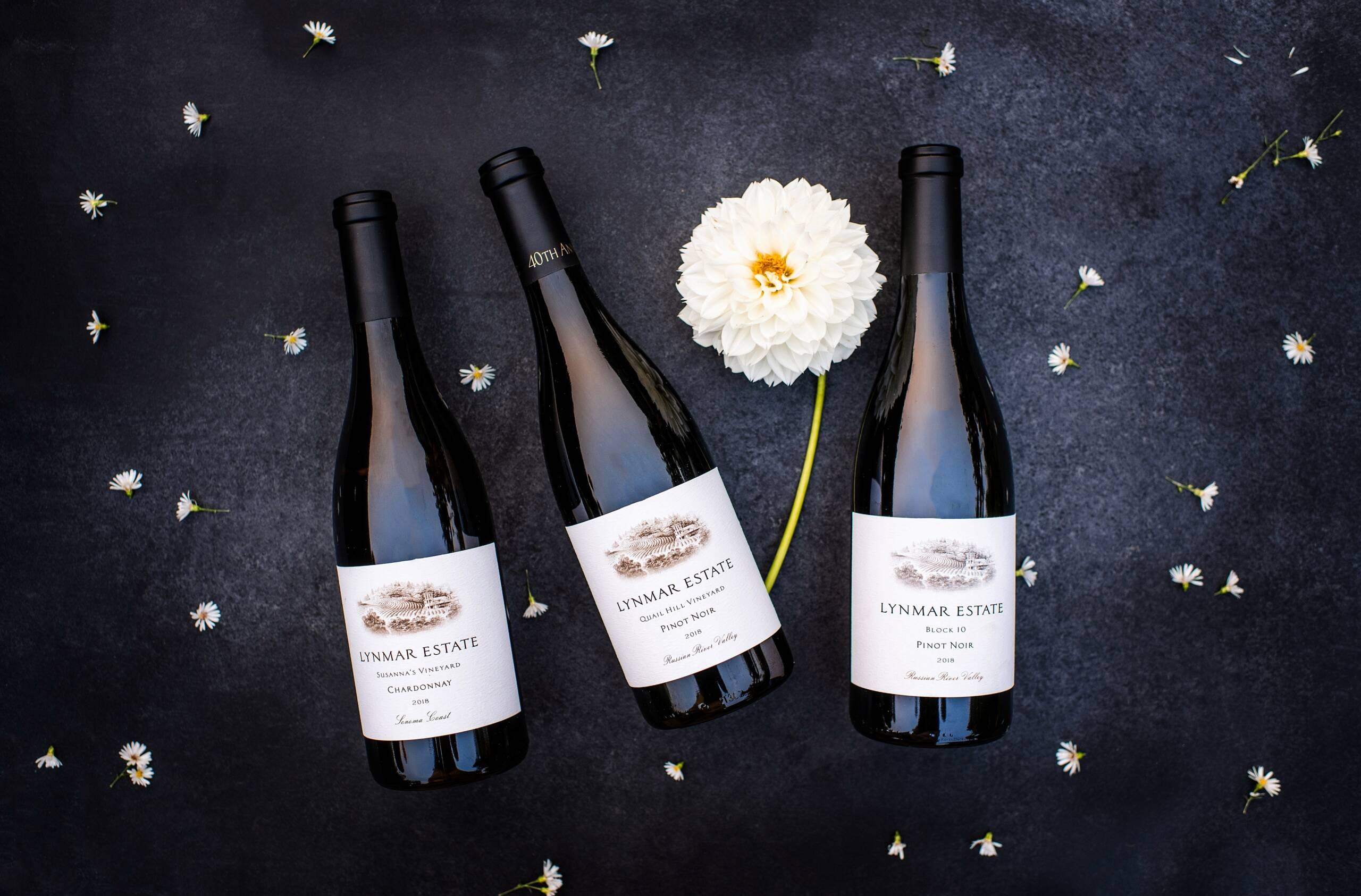 Three bottles of Lynmar Estate wine are arranged on a dark surface with scattered white flowers and a large white dahlia.