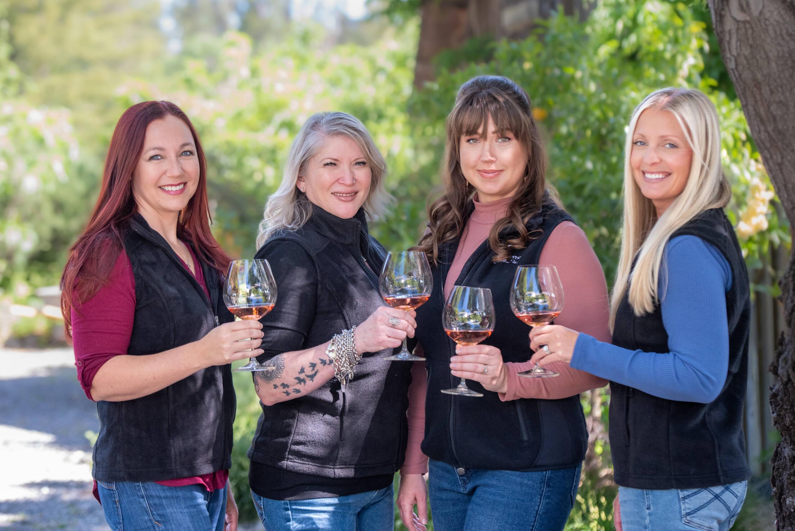 The Advocates Club team members pose outdoors, holding wine glasses and smiling at the camera. They are wearing casual clothes and vests, with greenery in the background.