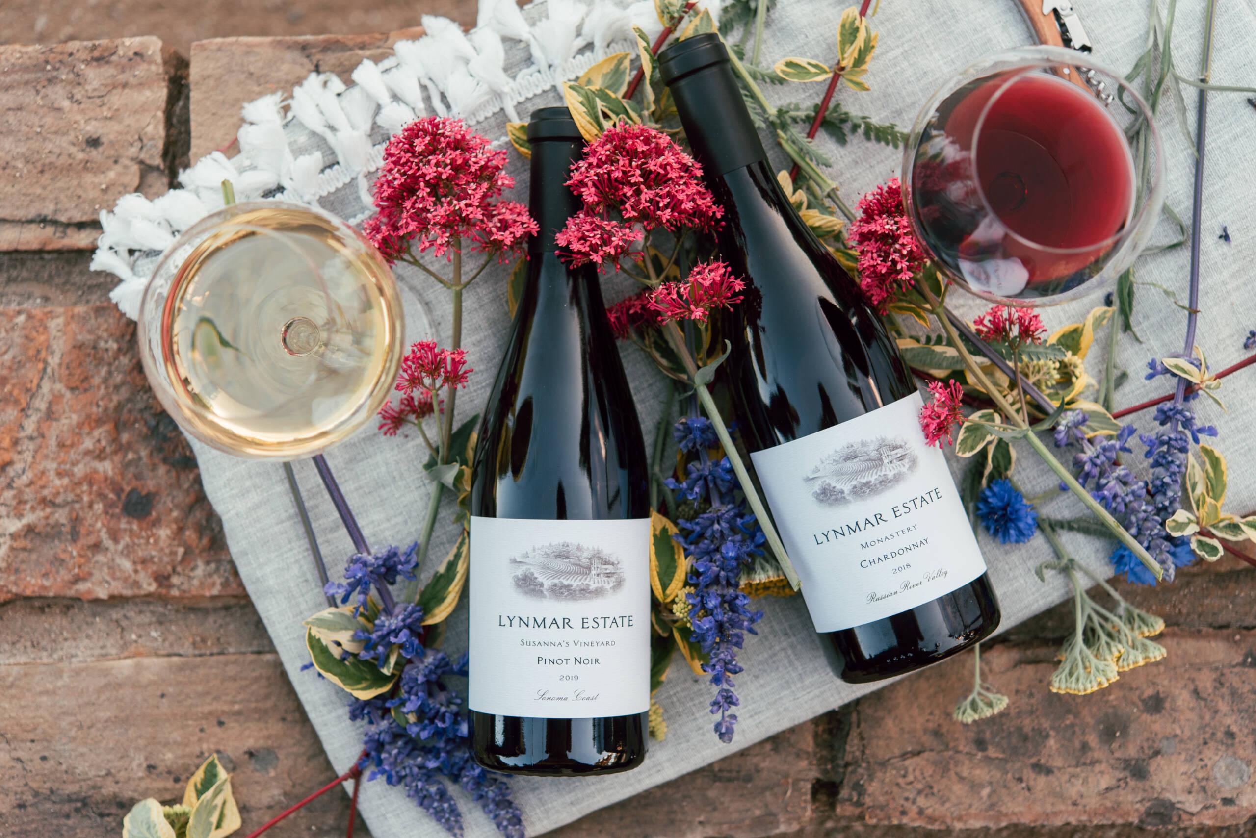 Two bottles of Lynmar Estate wine lie on a cloth surrounded by flowers, next to a glass of white wine and a glass of red wine on a brick surface.