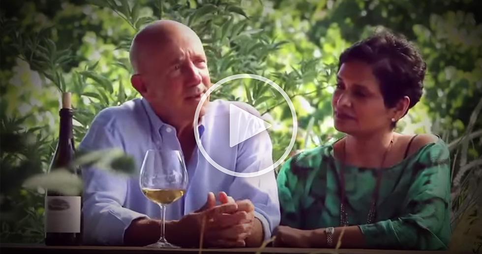 Lynn and Anisya Fritz sit outdoors at a table with a wine bottle and glass, surrounded by greenery.